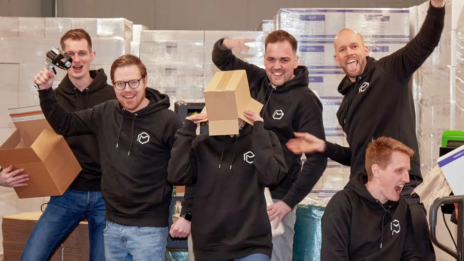 Six people from the Verpakgigant team, wearing black branded uniforms and blue jeans are playing around in the warehouse with boxes on their heads.