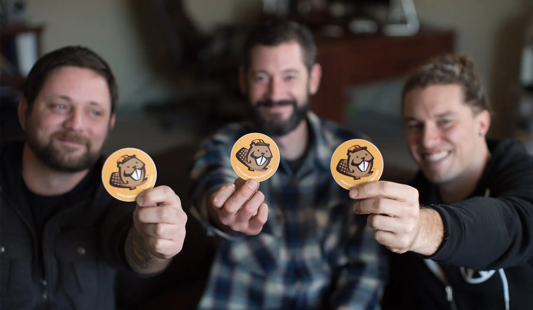 Three men from the Beaver Builder team hold up stickers of their beaver logo to the camera, with a shallow depth of field so their bodies are slightly out of focus.